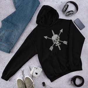 Puede incluir: Sudadera con capucha negra con un diseño de calavera gris con flechas cruzadas y una espada. La sudadera tiene un bolsillo delantero y una capucha. La imagen también muestra vaqueros azules, zapatillas deportivas, auriculares y un smartphone.