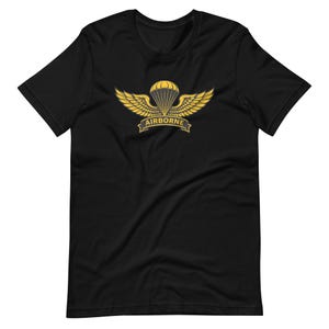 以下が含まれることがあります： 黒いTシャツ。金と黄色のAirborneの記章が特徴です。デザインにはパラシュート、翼、そしてバナーに「AIRBORNE」という文字が含まれています。このシャツは柔らかい素材でできており、半袖です。