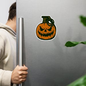 May include: A Halloween magnet depicting an orange pumpkin with a carved, sinister face, styled as a grenade. The top is green with a pull ring, outlined in white. The magnet is shown on a silver fridge.