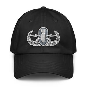 May include: A black baseball cap featuring an embroidered silver-toned Explosive Ordnance Disposal badge on the front. The emblem displays a central bomb shape, lightning bolts, and a surrounding laurel wreath, all stitched in shades of grey and silver.