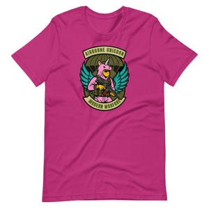 Könnte beinhalten: Magentafarbenes T-Shirt mit einem Aufdruck eines Einhorns in Militäruniform, das ein Gewehr hält. Das Design enthält den Text "AIRBORNE UNICORN" und "MODERN WARFARE" in einem Banner. Das Einhorn hat türkisfarbene Flügel und trägt einen rosa Overall.