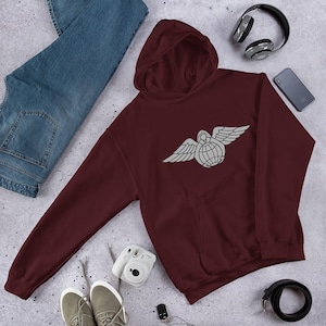 Può includere: Una felpa con cappuccio bordeaux con un emblema bianco di un globo alato. La felpa ha un cappuccio e una tasca frontale. Nell'immagine sono presenti anche jeans blu, scarpe da ginnastica grigie, una cintura nera, cuffie e uno smartphone.