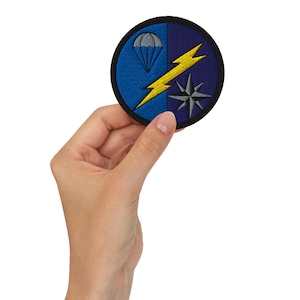 Op de afbeelding: Een ronde geborduurde patch met een gespleten blauwe en donkerblauwe achtergrond. Het ontwerp bevat een grijze parachute, een opvallende gele bliksemschicht en een grijs stervormig kompasembleem, alles omrand door een dikke zwarte rand.