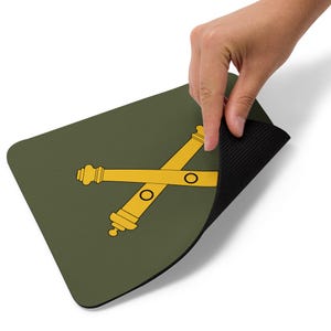 Könnte beinhalten: Ein rechteckiges, olivgrünes Mauspad mit einem goldenen, gekreuzten Kanonen-Design. Das Mauspad wird von einer Hand gehalten, wodurch die schwarze, strukturierte Unterseite sichtbar wird. Das Design umfasst zwei goldene Kanonen und zwei schwarze Kreise.