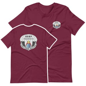 Könnte beinhalten: Burgunderfarbene T-Shirts mit kurzen Ärmeln und dem USAF Pararescue-Emblem auf der Vorder- und Rückseite. Das Emblem zeigt einen Fallschirm, einen Globus und den Text "That Others May Live."