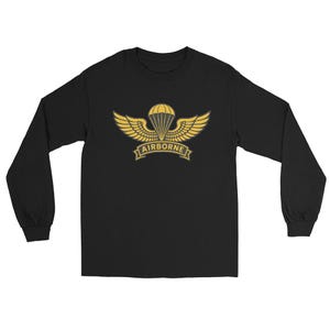 以下が含まれることがあります： 黒色の長袖Tシャツ。金色の空挺部隊のエンブレムが特徴です。エンブレムにはパラシュート、翼、そして「AIRBORNE」の文字がバナーに描かれています。このシャツは柔らかい素材で作られています。