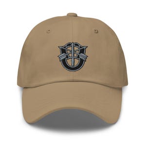 May include: A khaki baseball cap featuring an embroidered emblem on the front. The design consists of a black horseshoe shape with a silver-toned dagger pointing downward, flanked by two crossed silver-toned arrows, all centered on the crown.