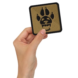Pode incluir: Patch quadrado com um desenho de pegada preta sobre um fundo bege, emoldurado por uma borda preta. A pegada tem um desenho de caveira no centro. O patch é segurado em uma mão.
