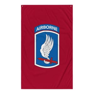 以下が含まれることがあります： 赤色の旗で、上部に「AIRBORNE」の文字が入った青と白のエンブレムが付いています。エンブレムには白い翼、赤い銃剣、青い背景が含まれています。