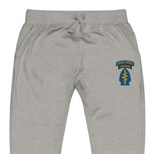 Peut inclure: Pantalon de survêtement gris clair avec une taille à cordon de serrage et des poches. La jambe droite comporte un écusson avec le texte "SPECIAL FORCES AIRBORNE" au-dessus d'un emblème bleu et jaune.