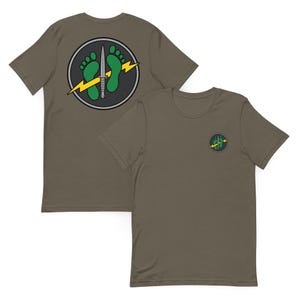 May include: An olive green short-sleeve t-shirt featuring a circular graphic on the front left chest and a larger version on the back. The design includes two green footprints, a silver dagger, and a yellow lightning bolt inside a dark circle.