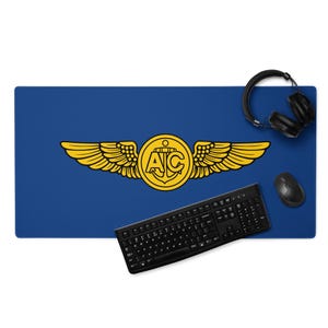 Könnte beinhalten: Blaues Schreibtisch-Pad mit einem goldenen Emblem mit Flügeln, einem Anker und den Buchstaben "AC". Eine schwarze Tastatur, Maus und Kopfhörer sind ebenfalls zu sehen. Das Pad ist rechteckig und für einen Arbeitsplatz konzipiert.