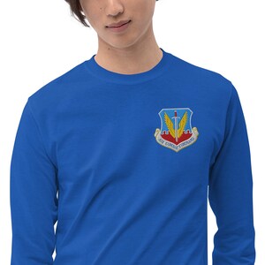 May include: Royal blue long-sleeve shirt with a patch on the upper left chest. The patch features a sword, wings, and the text "AIR COMBAT COMMAND". The shirt is made of a soft material.