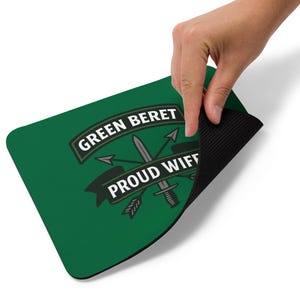 Puede incluir: Alfombrilla de ratón rectangular verde con el texto "GREEN BERET" y "PROUD WIFE" en blanco. El diseño incluye una espada, flechas y una daga. Una mano sostiene la alfombrilla.
