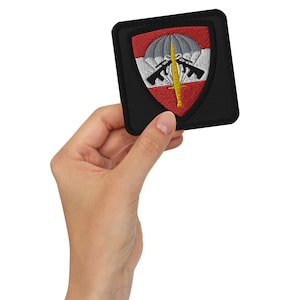 May include: A black embroidered patch with a red, white, and gray shield. The shield features a parachute, two crossed rifles, and a sword.