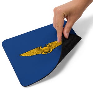 Könnte beinhalten: Blaues rechteckiges Mauspad mit einem goldfarbenen geflügelten Emblem. Das Emblem befindet sich mittig auf dem Pad. Das Mauspad hat eine schwarze, strukturierte Unterseite. Eine Hand hebt den Rand des Mauspads an.