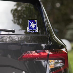 May include: A black car with a sticker on the back window. The sticker features a blue shield with a white knight riding a white horse. The text "NIGHT STALKERS" is below the shield.