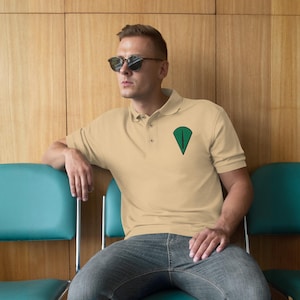 May include: A light tan polo shirt with a green and black emblem on the chest. The shirt has short sleeves and a button-down collar. The person is wearing sunglasses and blue jeans.