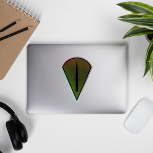 May include: A sticker featuring a black dagger design within a green, orange, and pink gradient shield shape. The sticker is placed on a silver laptop, with a notebook, pencils, headphones, a plant, and a mouse in the background.