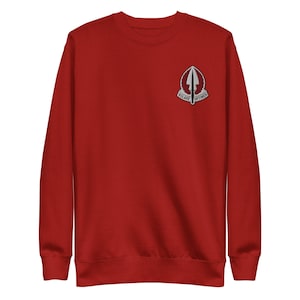 Pode incluir: Um moletom vermelho com gola redonda. Um pequeno emblema está na parte superior esquerda do peito. O emblema é um design branco e cinza com as palavras "VOLARE OPTIMOS".