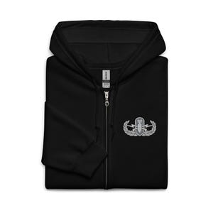 May include: A black zip-up hoodie featuring a hood with drawstrings and a full-length front zipper. On the left chest, there is an embroidered silver and grey military-style badge depicting a bomb, lightning bolts, and laurel leaf branches.