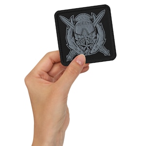 Op de afbeelding: Vierkante zwarte patch met een grijs geborduurd ontwerp. Het ontwerp toont een schedel met een bril, gekruiste zwaarden en een ster. De patch wordt in een hand gehouden.