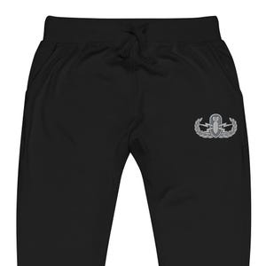 May include: Black sweatpants featuring an elastic waistband with a drawstring and an embroidered grey military-style explosive ordnance disposal badge on the right thigh. The badge depicts a bomb surrounded by lightning bolts and laurel leaves.