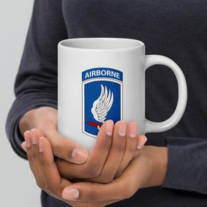 May include: White ceramic mug with a blue and white emblem. The emblem features the word "AIRBORNE" and a white wing design over a red and white shape. The mug has a large handle, perfect for tea or coffee.