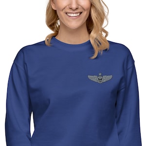 May include: A royal blue sweatshirt with a small embroidered emblem featuring wings, a star, and a circular design. The emblem is in shades of gray. The sweatshirt has long sleeves and a crew neck.