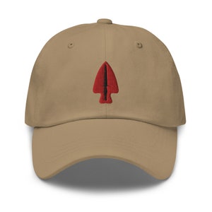 Puede incluir: Una gorra de béisbol de color beige con un diseño de punta de flecha bordado en rojo y negro.