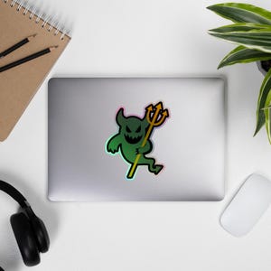 May include: A green devil sticker with a yellow trident. The sticker has a black face with horns and a neon outline. The sticker is on a laptop, with a notebook, pencils, headphones, a mouse, and a plant in the background.