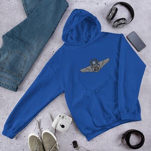 May include: A royal blue hoodie with a detailed emblem featuring wings, a star, and a circular design. The hoodie is laid flat, with a pair of blue jeans, sneakers, headphones, and a smartphone nearby. The hoodie has a drawstring hood and a kangaroo pocket.