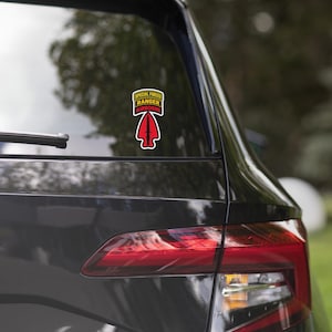 May include: A decal applied to a car window featuring a red arrowhead shape with a black dagger silhouette. Above the arrowhead are three stacked rectangular tabs with the text SPECIAL FORCES, RANGER, and AIRBORNE in yellow and red.