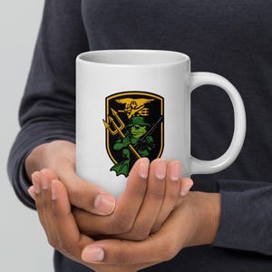 May include: White ceramic mug featuring a cartoon frog in a green hat and diving suit, holding a weapon and trident. The frog is inside a black and gold shield with an eagle and trident emblem. The mug has a large white handle.