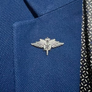 Può includere: Una spilla argentata USAF Special Warfare con ali, una spada e la scritta "USAF". La spilla è attaccata a un blazer blu strutturato. La fodera fantasia è visibile sul lato destro del blazer.