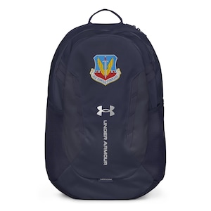 May include: A navy blue Under Armour backpack featuring the Air Combat Command emblem. The backpack has a top handle, side mesh pockets, and the Under Armour logo. The emblem is above the Under Armour logo.