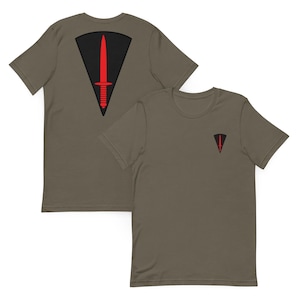 May include: An olive green t-shirt featuring a black triangular emblem with a red dagger graphic. The back of the shirt displays a large version of this design, while the front left chest area features a smaller, matching version of the same emblem.