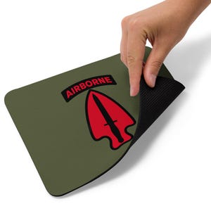 Op de afbeelding: Een olijfgroene rechthoekige muismat met een rood pijlpuntontwerp en een zwarte dolk, en het woord "AIRBORNE" in een rode rechthoek. De muismat heeft een zwarte, getextureerde onderkant.