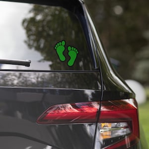 May include: Green baby footprint decal with a black outline, affixed to the rear window of a dark-colored vehicle. The decal is a simple, stylized representation of two footprints.