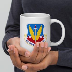 May include: White ceramic mug featuring the Air Combat Command emblem. The emblem includes a blue shield with a silver sword, yellow wings, and red and yellow accents. The words "AIR COMBAND" are printed on the mug.