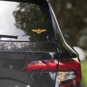 May include: A gold-colored sticker of a naval aviator's wings with a ship's anchor in the center, applied to the rear window of a black car.