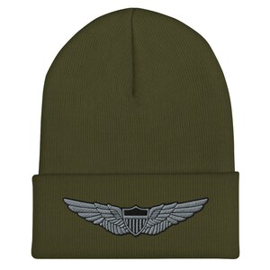 Pode incluir: Um gorro de malha verde com um remendo de asa de piloto bordado em prata. O remendo apresenta um escudo com uma estrela preta e uma borda prateada.