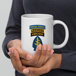 May include: White ceramic mug featuring the text "SPECIAL FORCES RANGER AIRBORNE" in blue, yellow, and black. Below the text is a blue and yellow emblem. The mug has a large handle.