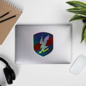 May include: A holographic sticker featuring a blue and red shield with a white eagle inside. The eagle is holding a yellow arrow in its talons.