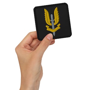 May include: A black embroidered patch with a gold winged dagger and a gold ribbon. The patch is held in a hand.