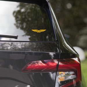 Può includere: Un adesivo dorato con un emblema alato è applicato sul lunotto posteriore di un veicolo grigio scuro. L'emblema presenta un design simmetrico con ali spiegate e una figura centrale, per scopi automobilistici o decorativi.