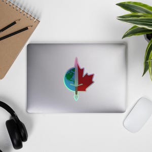 May include: A holographic sticker featuring a design that combines a red maple leaf, a globe, and a sword. The maple leaf is on the right, the globe on the left, and the sword runs vertically through the center. The sticker is on a laptop.