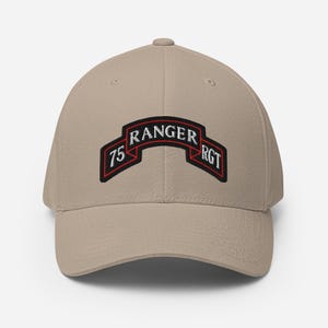 May include: A khaki baseball cap featuring an embroidered patch on the front. The patch is a black, arched banner with a red border and white text that reads 75 RANGER RGT. The cap has a structured crown, a curved brim, and stitched eyelets.
