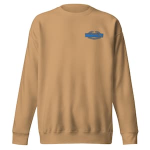 Può includere: Una felpa color beige con girocollo e maniche lunghe. Un emblema blu e grigio è ricamato sul petto in alto a sinistra. L'emblema presenta uno stendardo blu con testo e un design ad ala grigia.