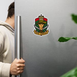 Pode incluir: Um íman colorido com o emblema dos Royal Gurkhas. O design inclui um escudo com uma coroa, um soldado com um chapéu, duas facas kukri cruzadas e o texto "Royal Gurkhas". O íman está preso a um frigorífico prateado.